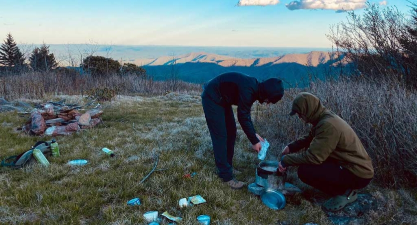Two people prepare food in an open grassy area. Beyond them are mountains. 